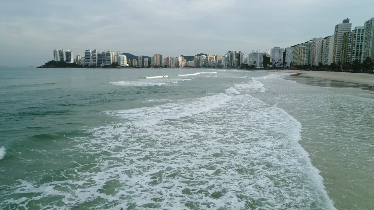 Toma de dron a baja altura pasando sobre pequeñas olas y mar en calma, con edificios al fondo en la ciudad, playa de Guarujá, Brasil.