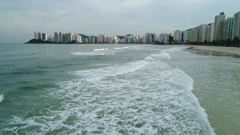 disparo de drones en plano bajo pasando sobre pequeñas olas y mar en calma, edificios en el fondo de la ciudad, guarujá, playa de brasil