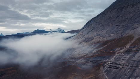 A-dark-mountain-towering-above-the-landscape,-autumn-forest-covers-the-foothills,-white-fog-floating-in-the-air