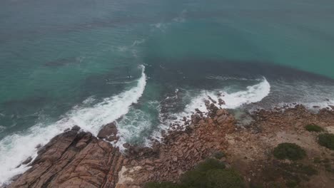 Group-of-surfers-floating-close-to-rocky-coast-in-turquoise-water-sea-aerial-high-shot-in-Margaret-river,-Australia