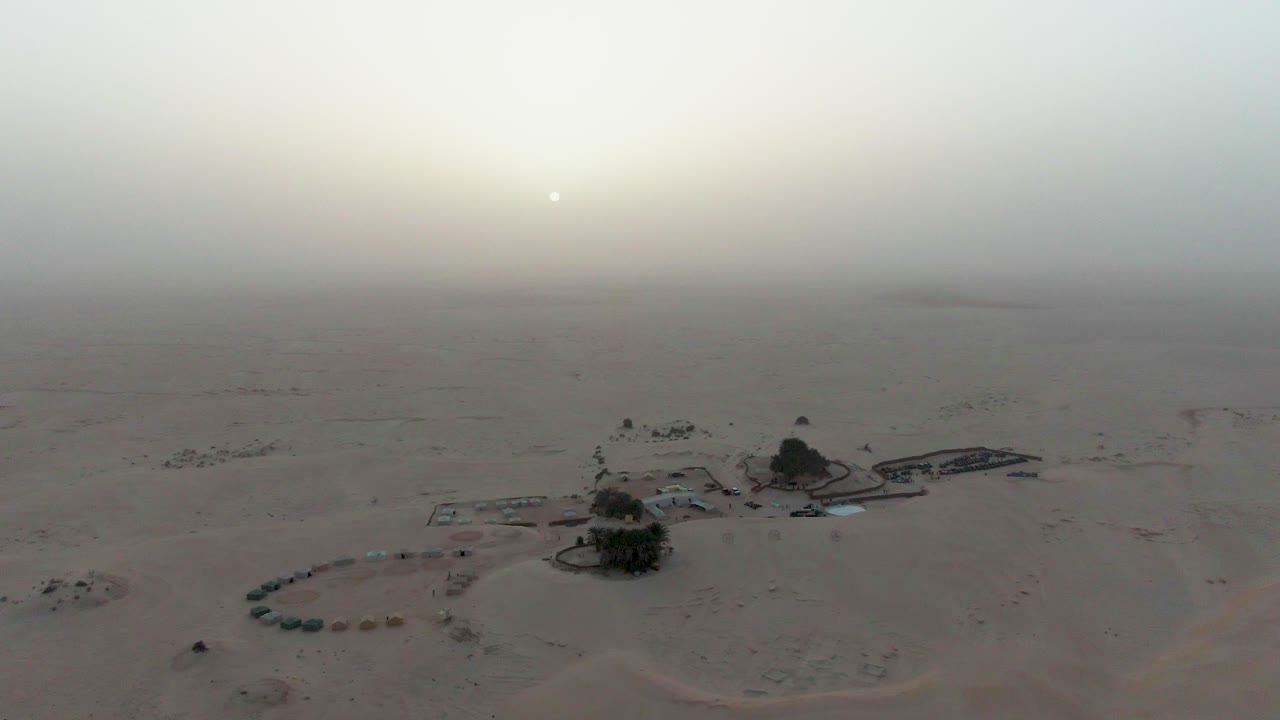 Vista aerea di un campo desertico isolato all'alba, con una leggera foschia che circonda piccole strutture e alberi, creando una sensazione di isolamento, silenzio e vita senza tempo in un paesaggio sabbioso infinito.