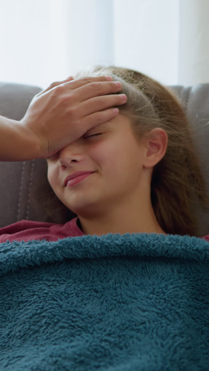Niña pequeña durmiendo arropada con una manta, sonriendo cálidamente, mientras una persona le toma la temperatura. Luz natural entrando por la ventana.