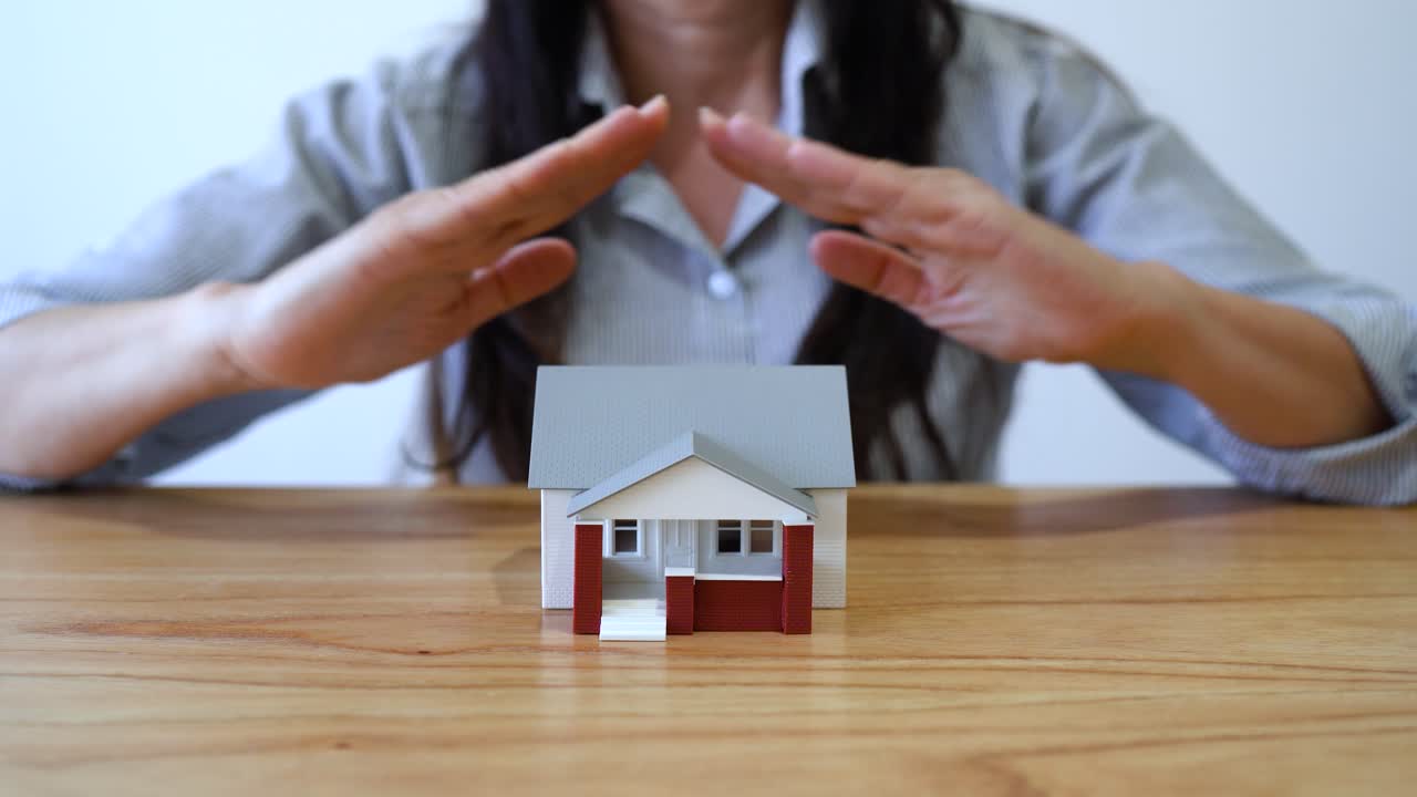 Woman Places Hands In Roof-like Gesture Over Model House On Table. House Insurance Concept. closeup shot