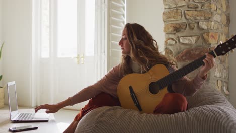 caucasian woman sitting on beanbag playing acoustic guitar using laptop in sunny cottage living room