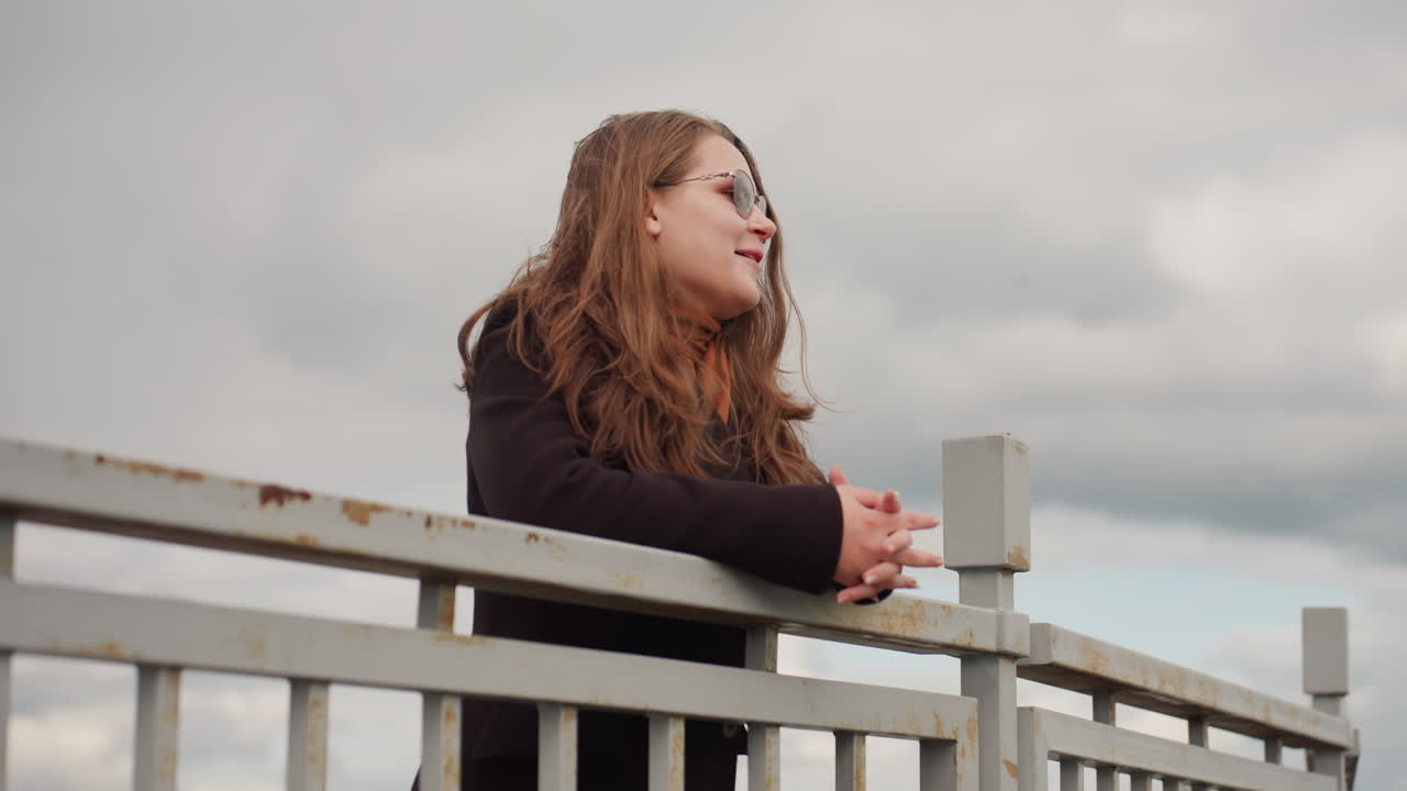 Charming woman with long hair wearing eyeglasses turns gracefully giving wide smile as gentle breeze moves her hair, creating natural warmth and expression of confidence and joy outdoors