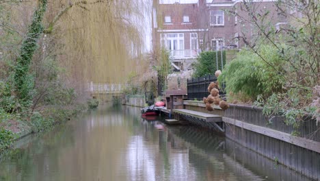 Calm-treelined-stretch-of-canal-in-Utrecht,-Netherlands,-with-reflections-on-the-water-and-traffic-driving-over-a-bridge-in-the-distance