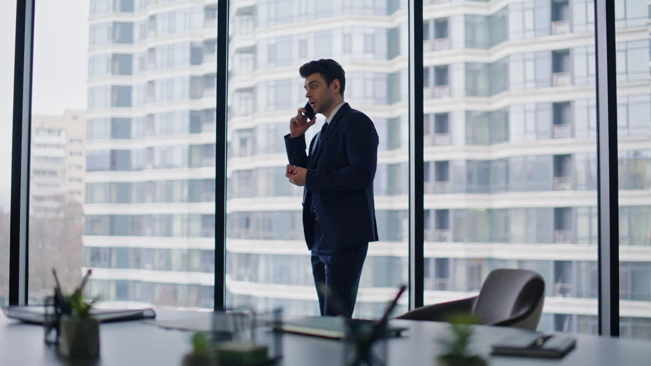 Investitore di successo parla al cellulare discutendo un accordo vicino alla finestra di un ufficio