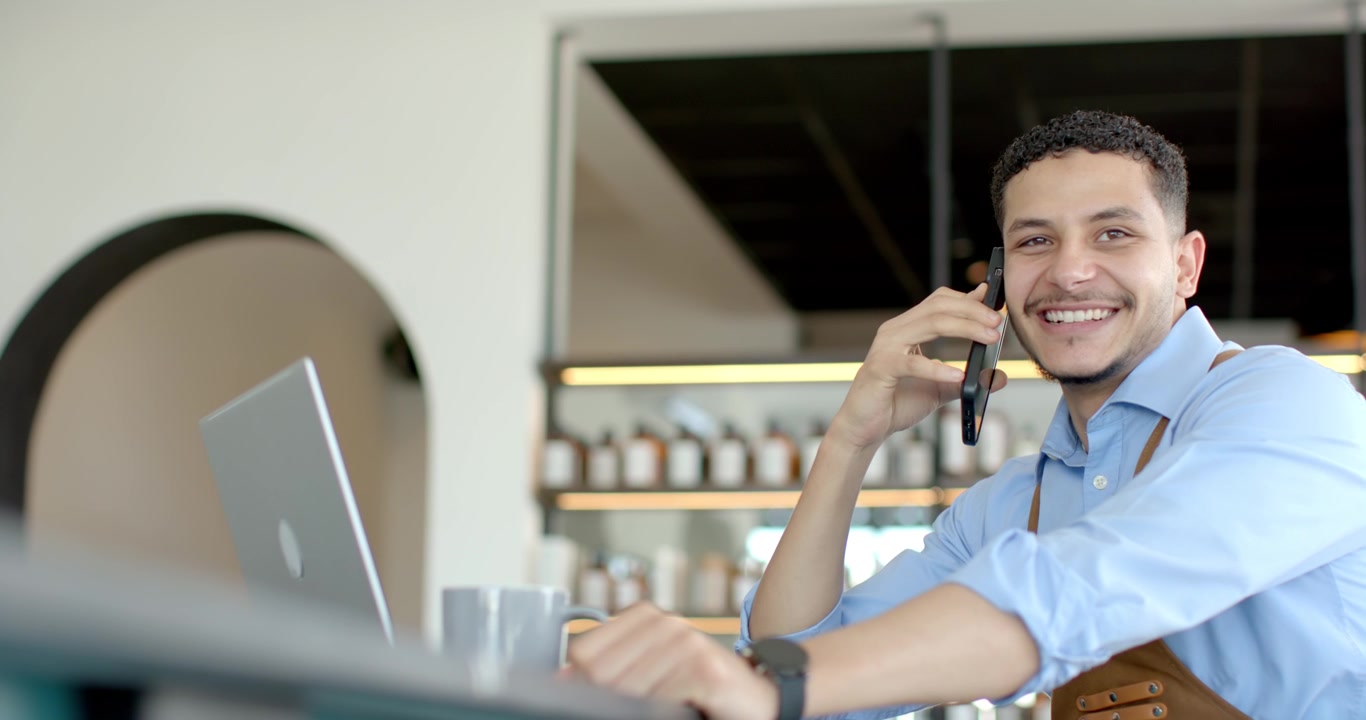 Hombre africano con delantal de cuero contestando una llamada, sosteniendo un teléfono y alcanzando una taza en el mostrador de una cafetería