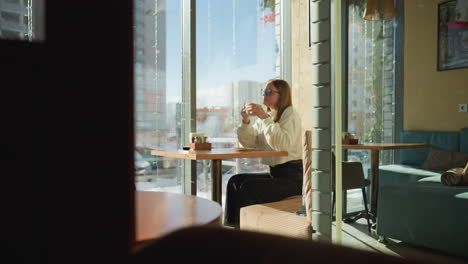 girl seated at table sipping tea in cozy cafe with view of snowy street, residential buildings, and parked cars outside, soft sunlight and indoor decor create warm, relaxed winter atmosphere