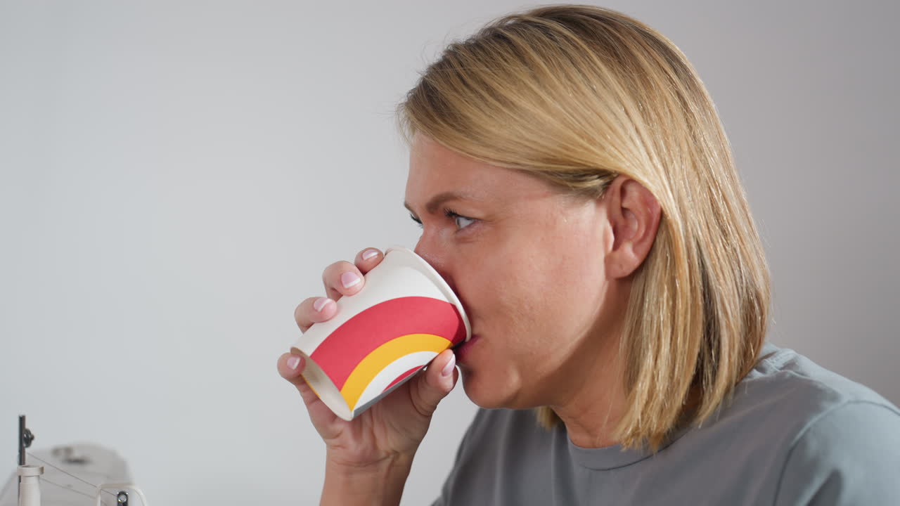 Female seamstress seated beside sewing machine in bright tailoring workspace enjoying drink and biscuit snack while communicating with fellow seamstress, sharing friendly moment during break
