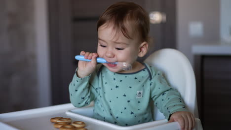 one-year old toddler sitting at the feeding table with spoon in mouth. child turns to bagels left in front of him. blurred background.