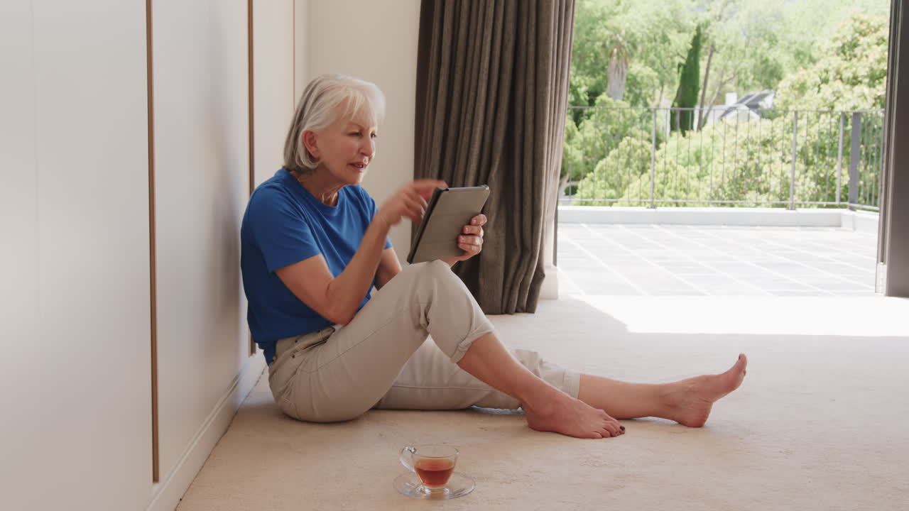 Senior woman relaxing at home, using tablet with cup of tea nearby