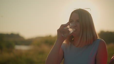 mujer de camiseta azul bebe un refrescante vaso de agua al aire libre, rodeada de verde sereno y bañada en la cálida luz del sol, disfruta de un momento de paz en la naturaleza, con un fondo escénico borroso