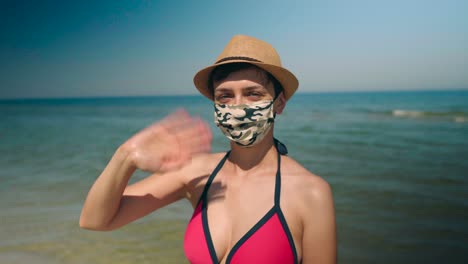 Mujer-Joven-En-Bikini-En-La-Playa-Con-Mascarilla-Y-Sombrero-Para-El-Sol-Saludando