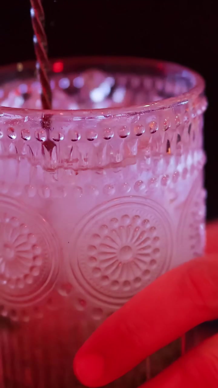 Barman mescola un cocktail con ghiaccio in un bicchiere decorato sotto vivaci luci del bar, primo piano.
