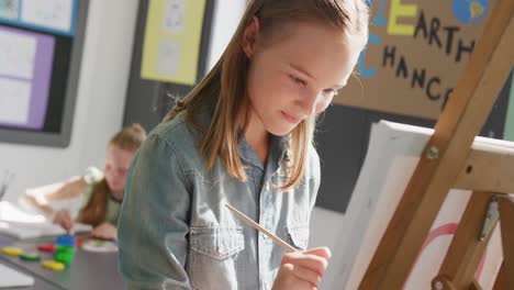 video of happy caucasian schoolgirl standing at easel painting in art class, copy space