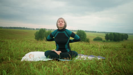 old woman sitting on mat in sukhasana pose with eyes closed, practicing yoga in a vast grassy field under a cloudy sky, with trees lined up in the background