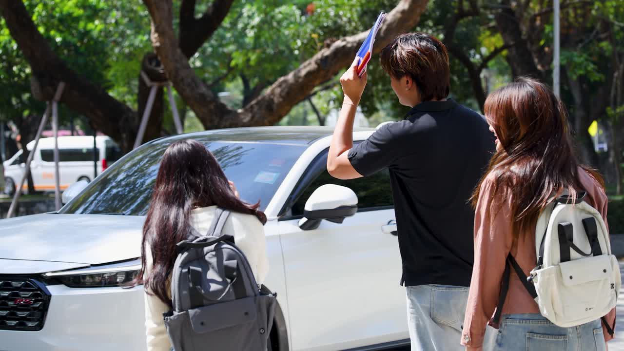 Trois étudiants thaïlandais discutent et rient ensemble à l’extérieur, sur un campus universitaire ensoleillé avec des voitures