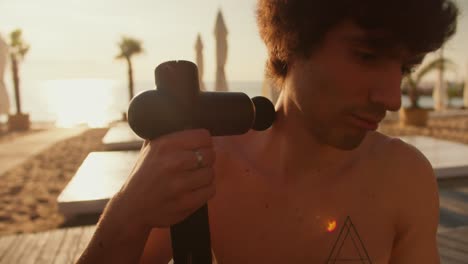 the guy massages his neck with a special vibrating massager. relaxation after morning workout on the beach at sunrise