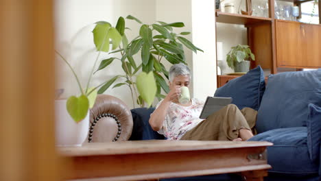 Happy-senior-biracial-woman-sitting-on-couch-drinking-tea-and-using-tablet-at-home,-slow-motion