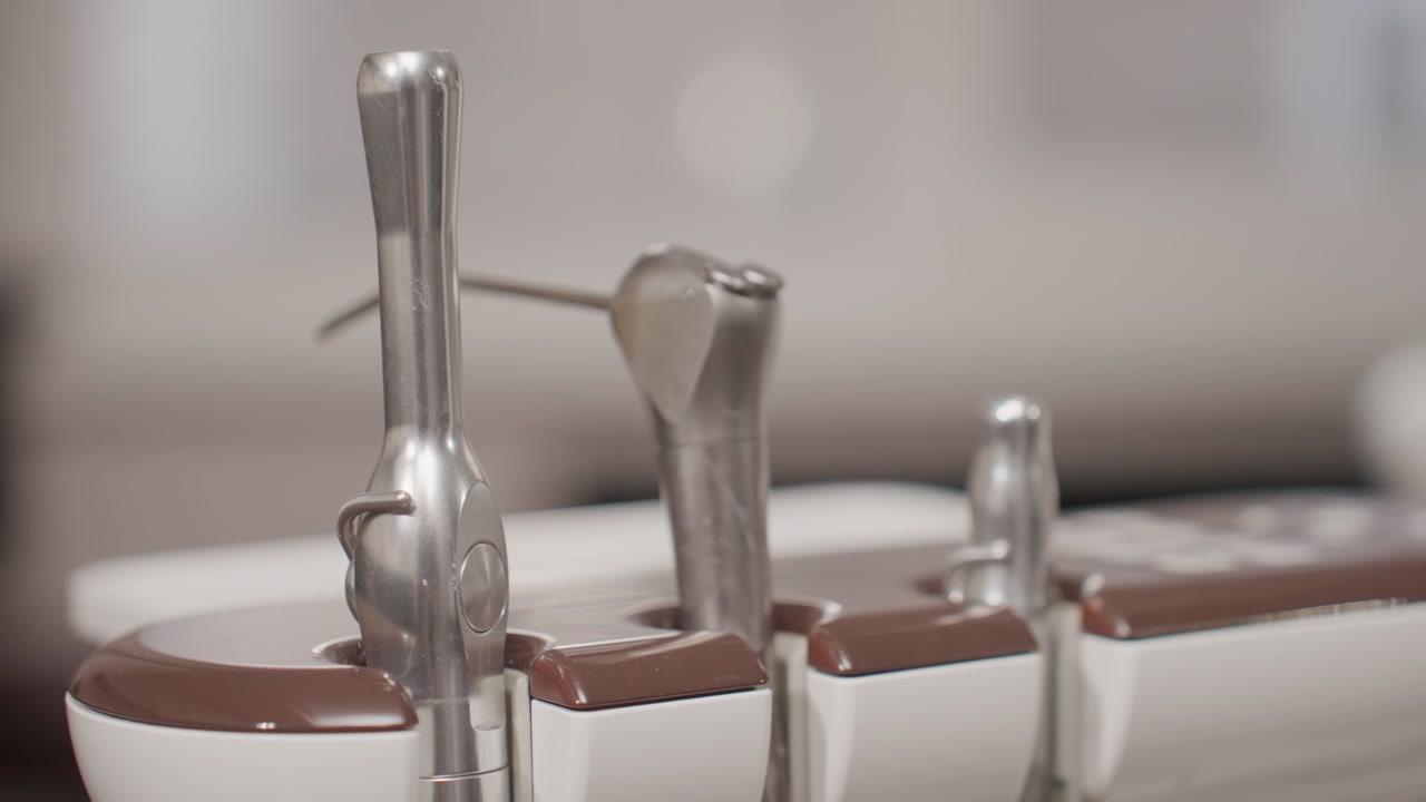 Close-up de peças de mão dentárias em seus suportes, exibindo ferramentas metálicas e recipientes brancos com tampas marrons. Iluminação suave realça superfícies lisas sem sombras fortes, com foco.