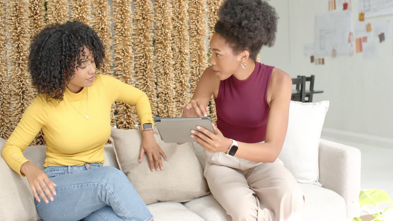 Diverse women sitting on sofa, one in mustard top, right woman showing tablet content, left tapping