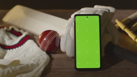 teléfono móvil de pantalla verde rodeado de pelota de bate de cricket y ropa en superficie de madera 2