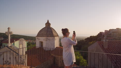 Glückliche-Frau,-Die-Mit-Ihrem-Smartphone-Ein-Foto-Des-Wunderschönen-Sonnenuntergangs-Macht-Und-Es-Genießt,-Ihr-Sommerurlaubsreiseerlebnis-Auf-Dem-Balkon-Zu-Teilen