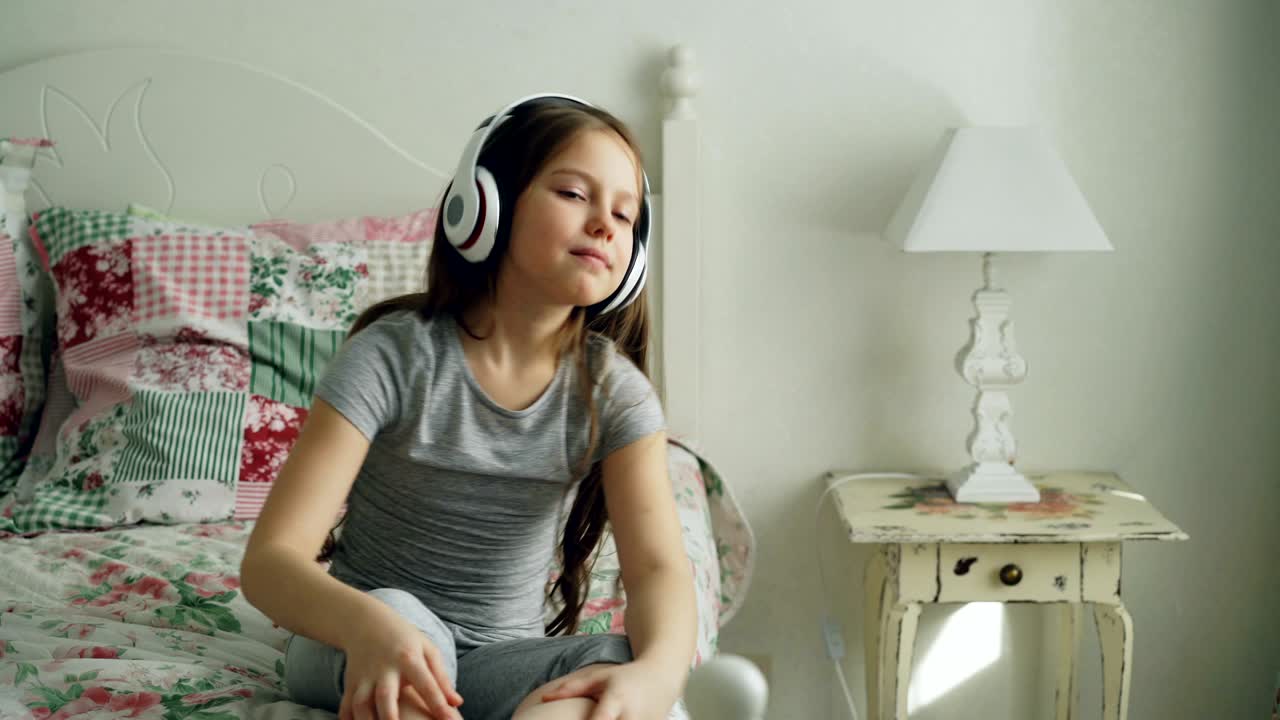 헤드폰을 입고 미소 짓는 귀여운 어린 소녀가 춤을 추고 머리를 웃기게 움직여서 집에서 침대에 앉아서 편안한 침실에서