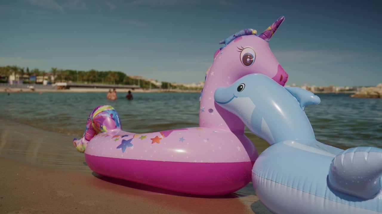 Colorful unicorn and dolphin inflatable floats on the shallow water at Plage de la Gravette with families swimming and the rocky breakwater in the background