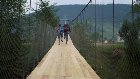 Friends-run-racing-outdoors-on-mountains-bridge.-Tourists-wear-backpacks-on-hike