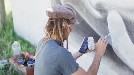 Video-Eines-Kaukasischen-Künstlers-Mit-Dreadlocks,-Der-Sein-Smartphone-Benutzt-Und-Ein-Walwandbild-An-Die-Wand-Malt