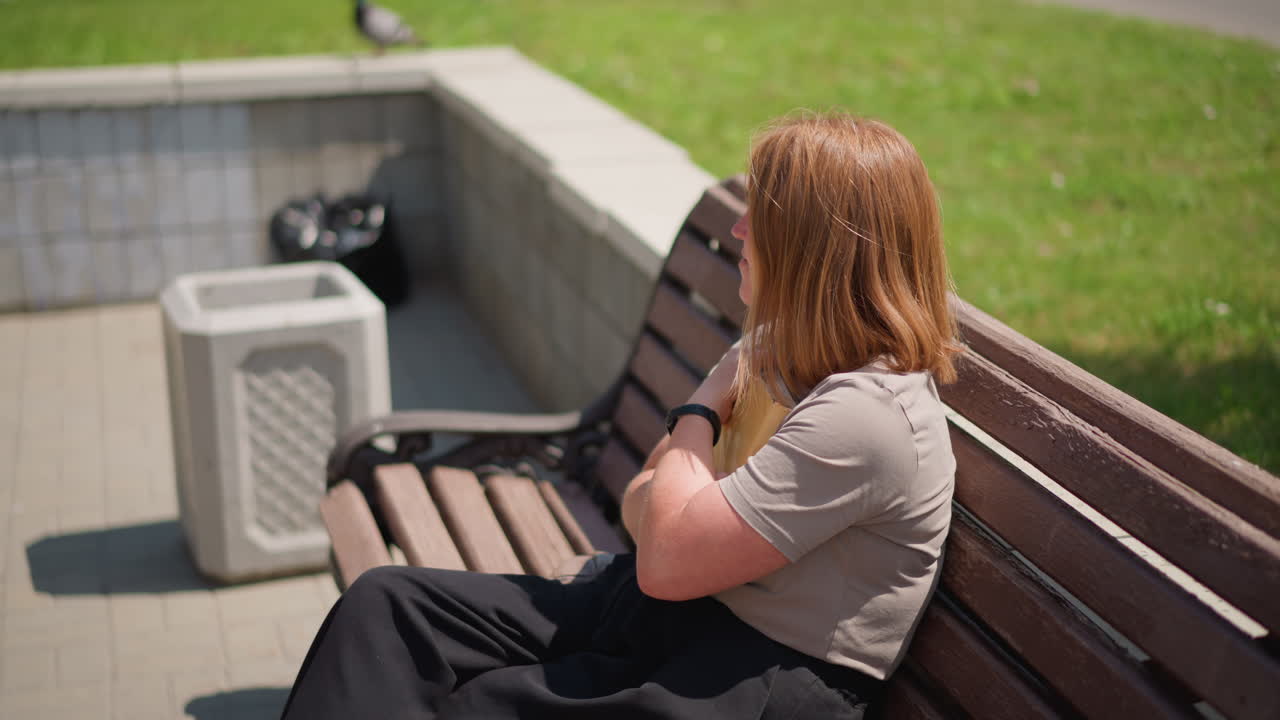Calm lady sits on wooden bench under warm sunlight flipping book pages then gently closes it and presses it to chest while pigeon flies nearby surrounded by green trees