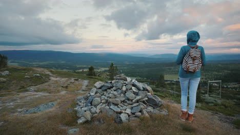 A-Woman-Is-Standing-At-A-Pile-Of-Stones-On-A-Mountain-In-The-Background-Of-A-Dramatic-Sky-A-Rear-Vie