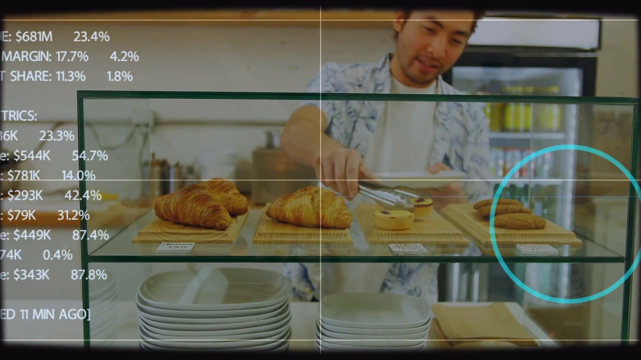 Un employé de boulangerie repère une pâtisserie, la saisit avec des pinces et la dispose pour la vente, avec une interface HUD superposée à la vitrine.