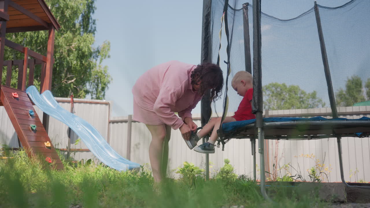 Woman Fastening Child Shoe By Trampoline, Warm Family Moment In Grassy Backyard With Slide And Netted Trampoline Attentive Caregiver Ties Laces While Toddler Waits, Gentle Focus And Safe Playful