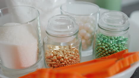 close-up of diverse colorful beads in gold, green, and pearl shades stored in glass jar, sugar in glass cup and orange baking tools arranged neatly in white bowl