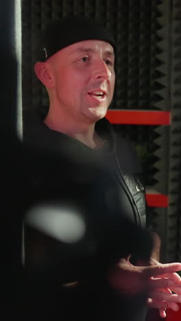 un hombre con una camisa negra y una gorra habla con entusiasmo con otra persona, apenas visible, en contra de una habitación negra a prueba de sonido con llamativos detalles rojos, destacando un momento de comunicación vibrante
