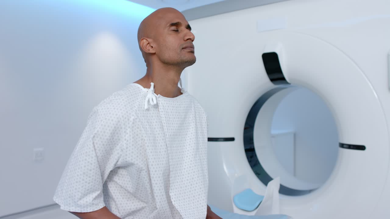 Uomo afroamericano in camice vicino a uno scanner CT, che rilassa le mani, solleva la testa e chiude gli occhi in attesa della scansione.