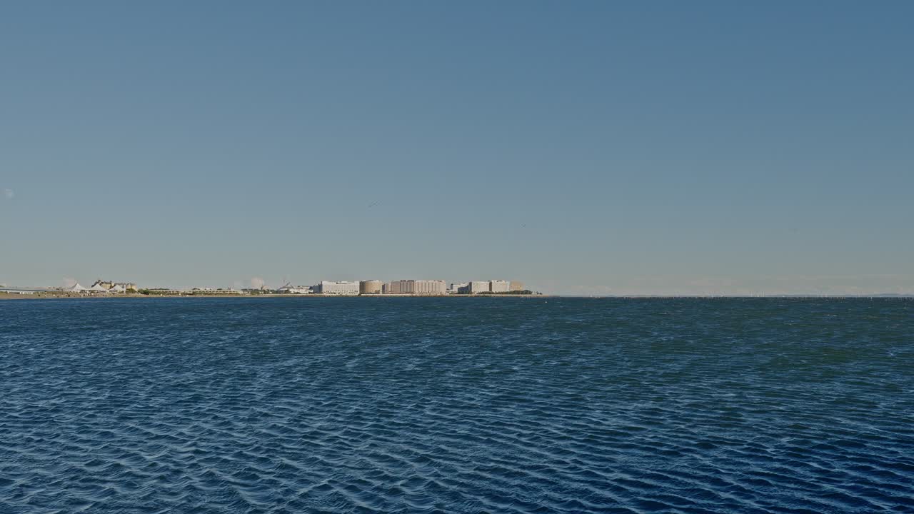 A very wide shot of blue water dominating the frame with a cluster of low-rise buildings on the distant horizon under a clear sky