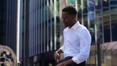 Tired-Young-Businessman-In-Shirt-Sleeves-Looking-At-Mobile-Phone-Standing-Outside-Offices-In-The-Financial-District-Of-The-City-Of-London-UK