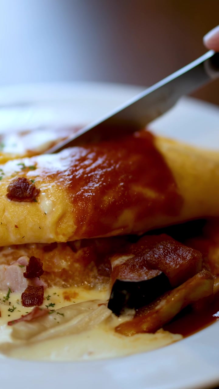Un coltello taglia l'omurice su un piatto bianco, rivelando il ripieno, sotto luce naturale in un ambiente da ristorante