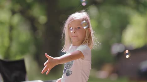 Una-Chica-Rubia-De-Ojos-Brillantes-Disfruta-De-Un-Día-Soleado-Al-Aire-Libre,-Mientras-Sopla-Delicadamente-Pompas-De-Jabón-Que-Brillan-Y-Flotan-A-Su-Alrededor.