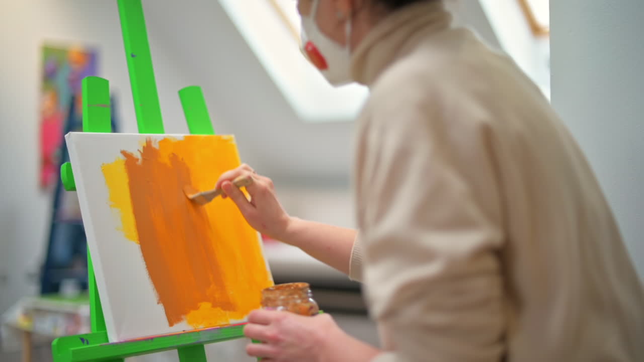 A woman in medical mask painting a picture using a brush in a studio. Slow motion