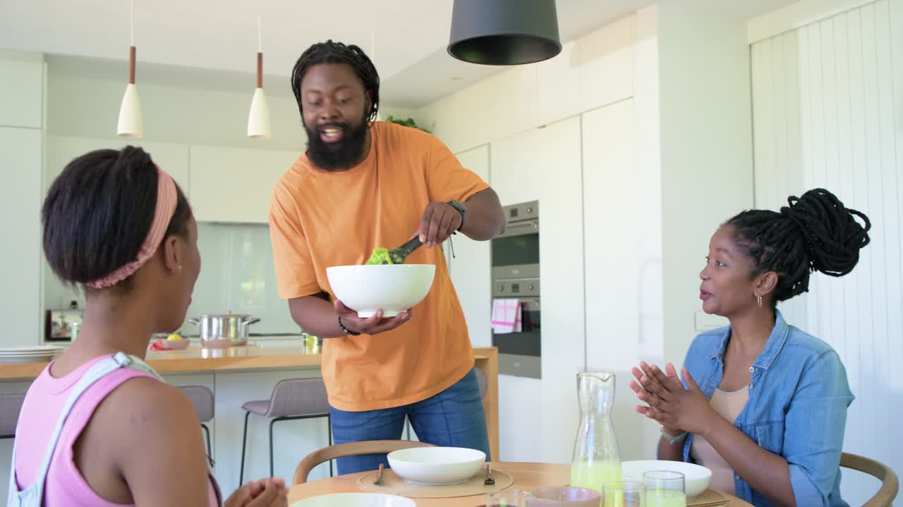 Adult man responding to women's clap, serving salad from white bowl at dining table for family