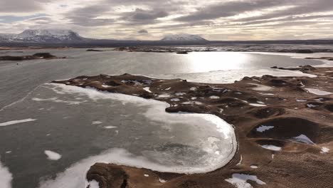 aerial view of the partially frozen lake mývatn, iceland, with volcanic formations and distant snowy mountains under a cloudy sky.
