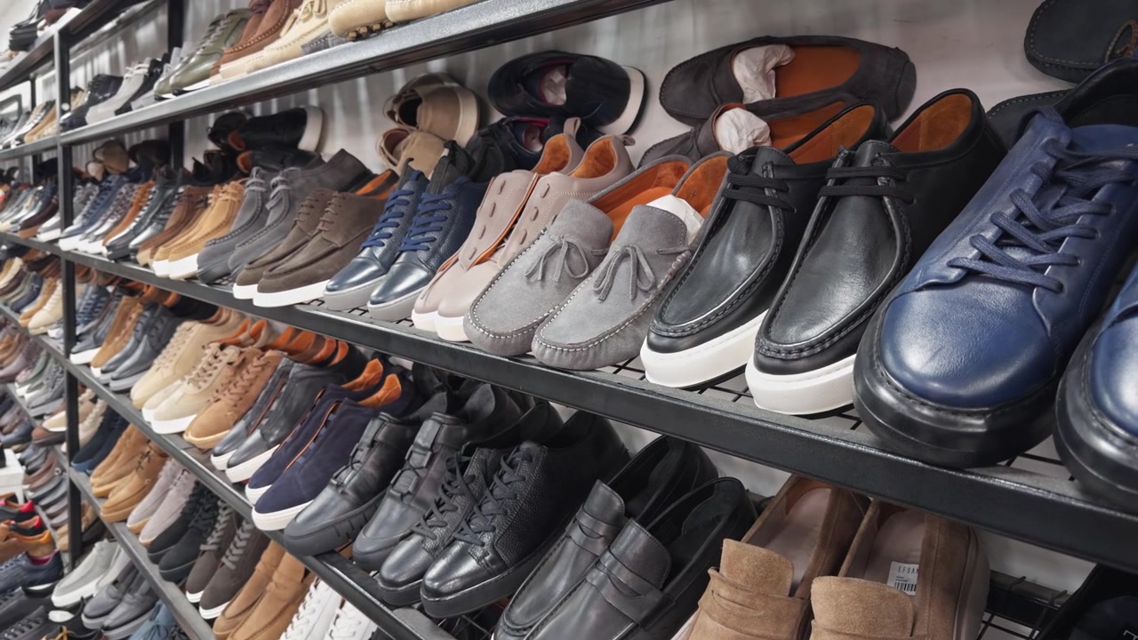 Kinoreifes 4K-Filmmaterial von Herrenschuhen, die ordentlich in Metallregalen in einem Einzelhandelsgeschäft angeordnet sind. Loafer, Oxfords, Sneaker und Mokassins in verschiedenen Farben und Materialien sind ausgestellt.