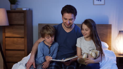 Padre-Guapo-Leyendo-Un-Libro-De-Cuentos-De-Hadas-Para-Su-Pequeño-Hijo-Y-Su-Hija-Antes-De-Irse-A-Dormir-En-El-Dormitorio-Por-La-Noche