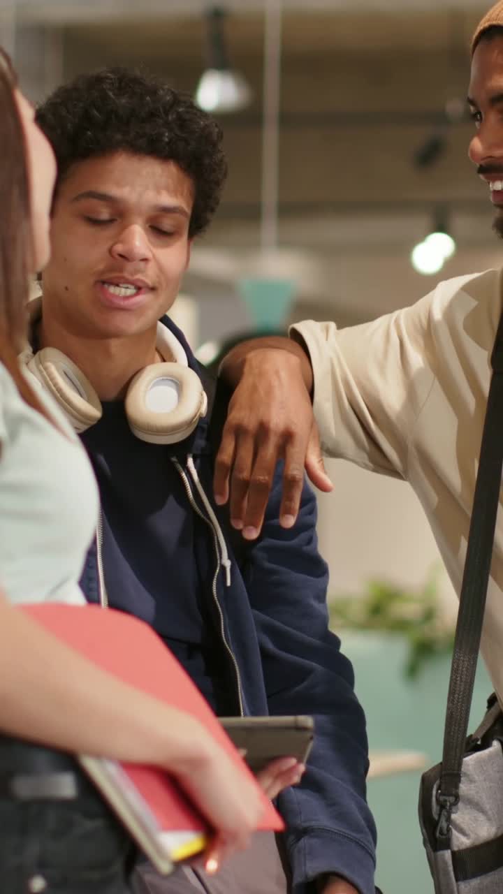 Vertical video: Leaning beanie man greeting hoodie in campus lounge, phone red notes woman joining