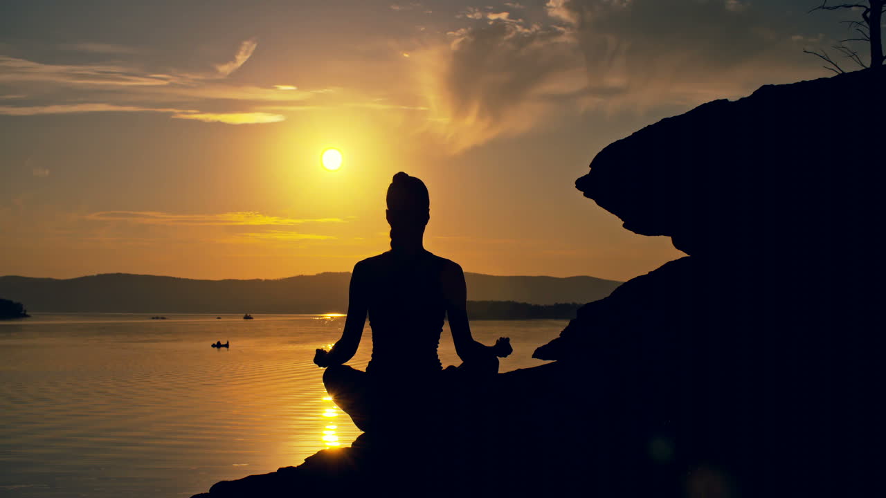 vista retroiluminada de una mujer haciendo poses de yoga sentada en una piedra frente al mar al atardecer